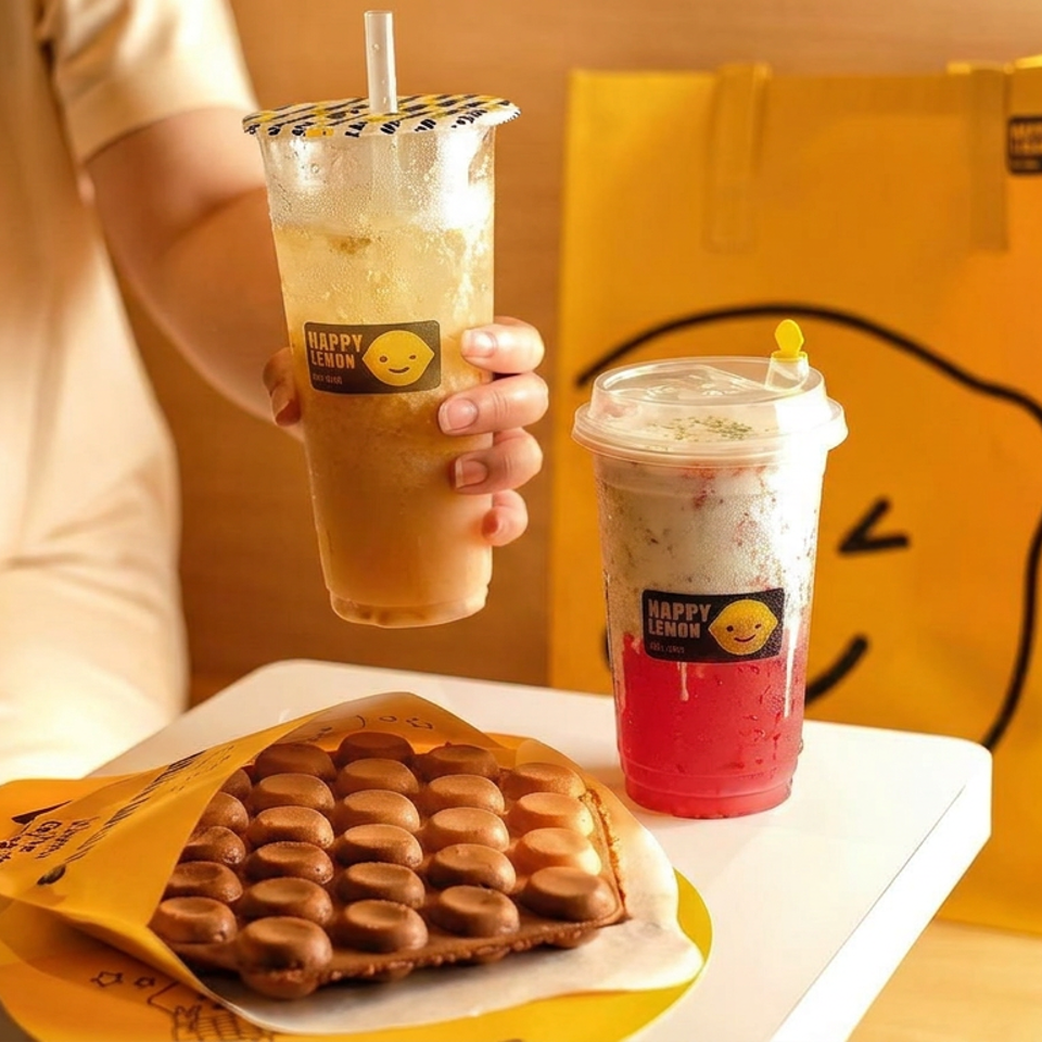 Person holds a lemon drink beside a red beverage and a golden bubble waffle on branded yellow packaging.