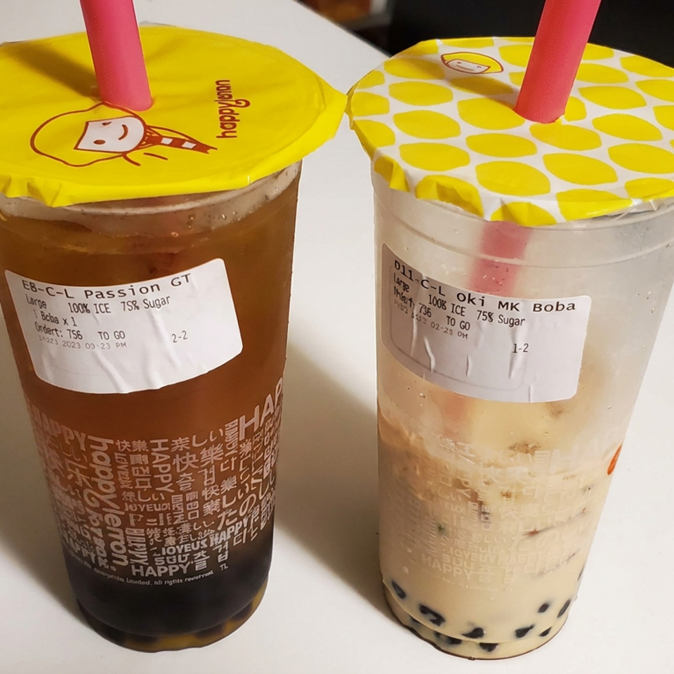 Two Happy Lemon bubble teas on a table: passion fruit green tea and Okinawa milk tea with tapioca pearls, yellow lids, and pink straws.