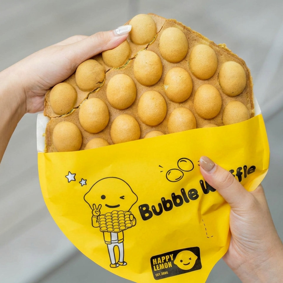Hands hold a golden bubble waffle in bright yellow Happy Lemon packaging, showing its distinctive round bubbles and cartoon branding.