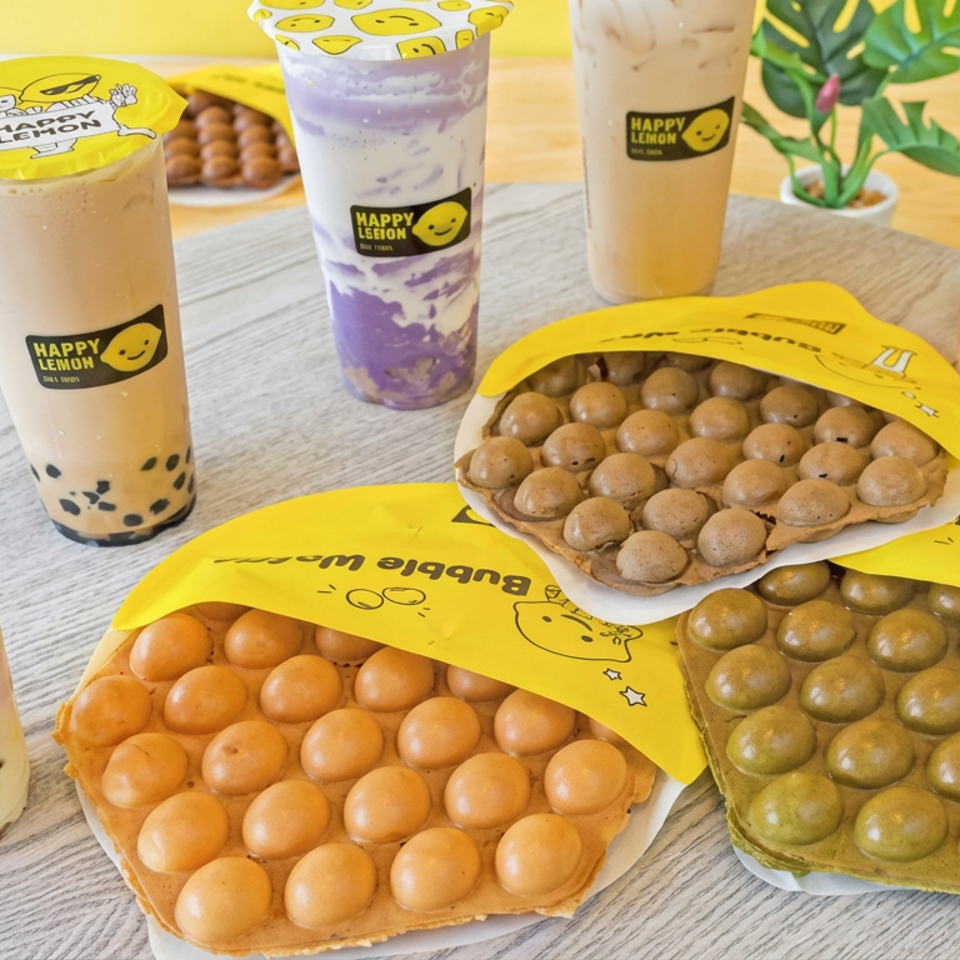 Five colorful Happy Lemon drinks and three orange, green, and chocolate bubble waffles arranged on a wooden table.