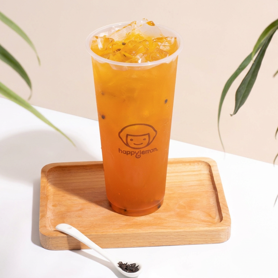 Orange passion fruit drink with ice in a branded cup, presented on a wooden tray amid green plants.